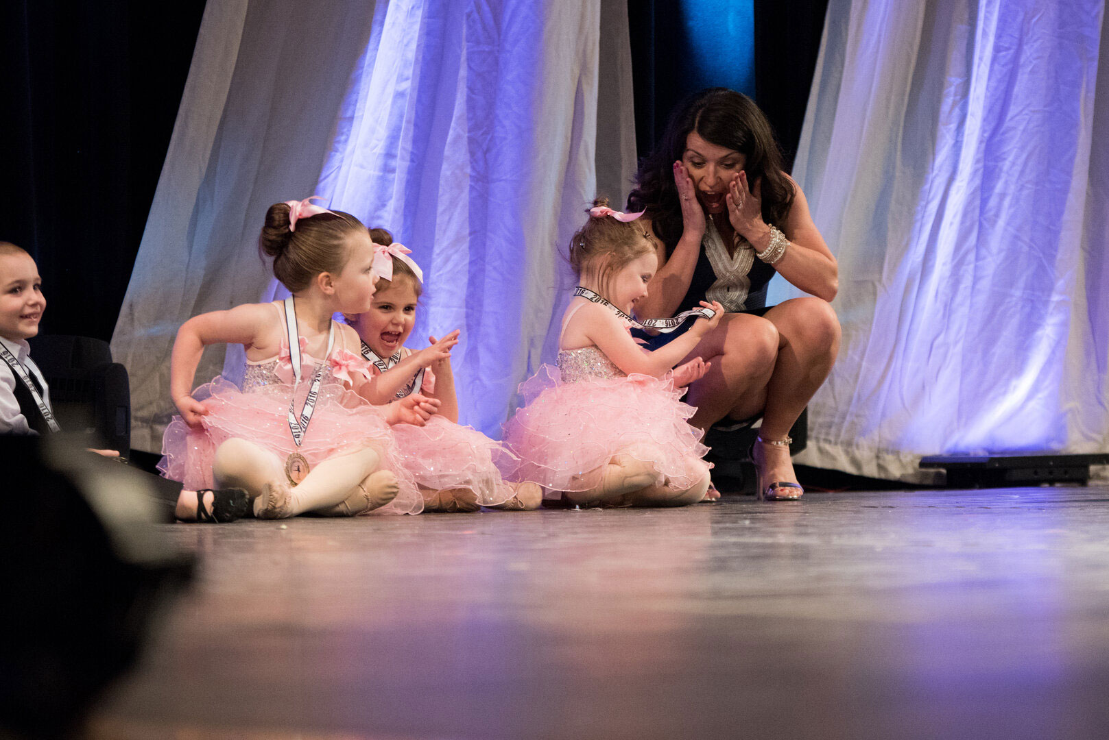 Young girls in tutu costumes sitting on stage with a woman.