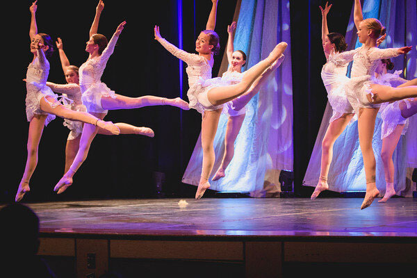 Three ballet dancers perform a jump on stage in white costumes.